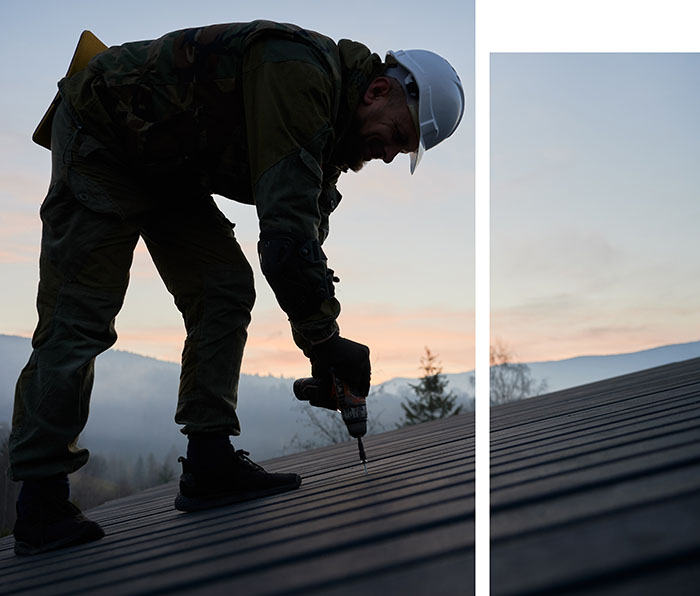 A worker on a rooftop, wearing a safety helmet, uses a drill during sunrise, surrounded by misty mountains and trees.