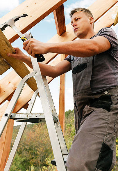 A person stands on a ladder holding a hammer, preparing to work on wooden beams of a partially constructed roof.