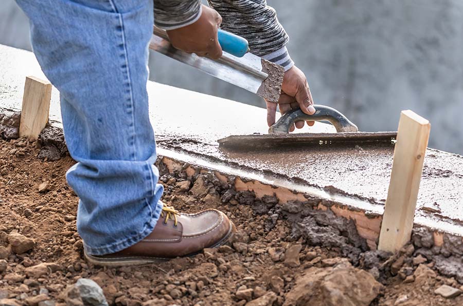 A worker smoothing freshly poured concrete with a trowel, wearing jeans and boots, beside wooden stakes on a dirt surface.