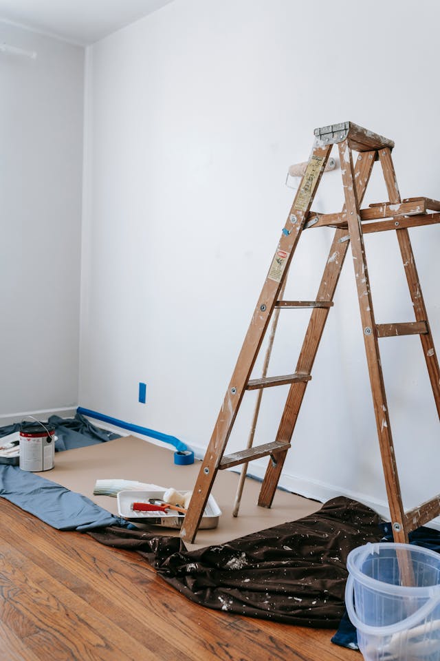 A wooden ladder stands next to a freshly painted white wall, surrounded by paint supplies and drop cloths on a hardwood floor.