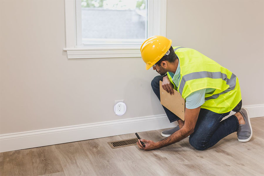 A construction worker in a hard hat and reflective vest kneels by a floor vent, taking notes on a clipboard in a light-colored room.