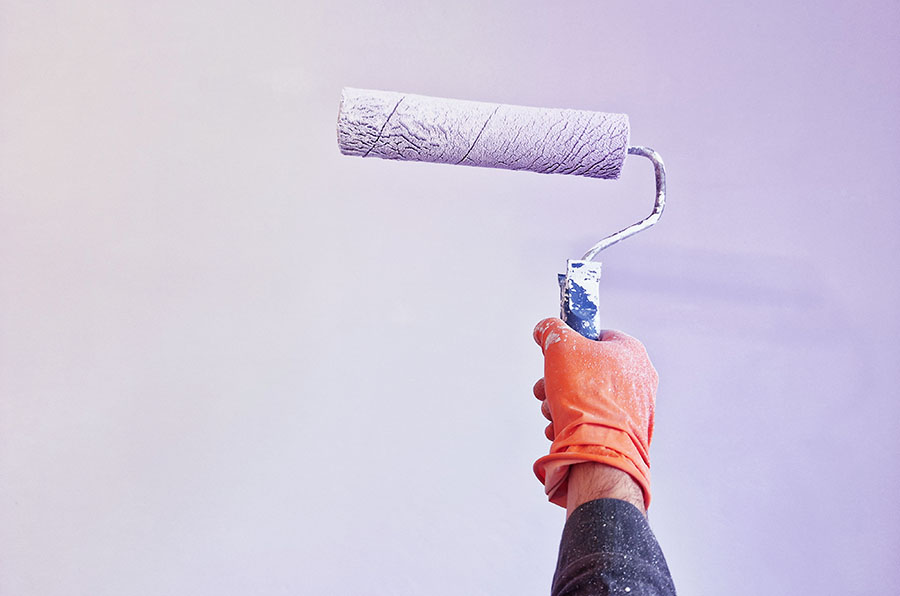 A hand wearing an orange glove holds a paint roller against a light purple wall, ready for painting.