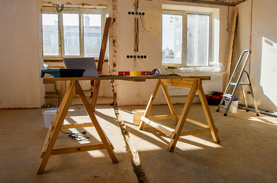 A wooden work table with a laptop, tools, and a ladder in a sunlit, unfinished room with bare walls and exposed wiring.