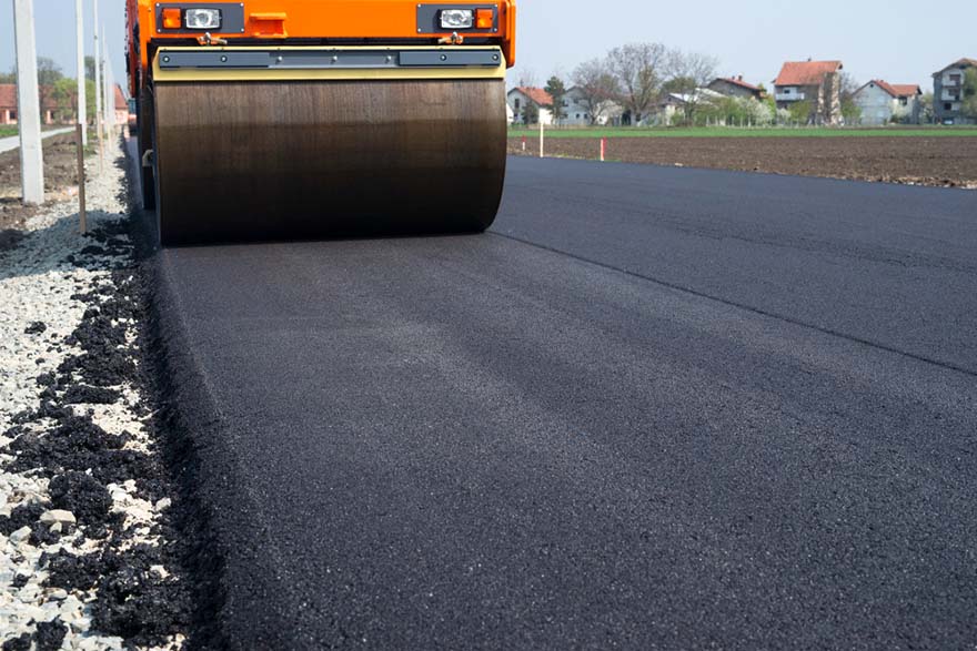 A large road roller smooths freshly laid asphalt on a new roadway, with houses and farmland in the background under a clear blue sky.